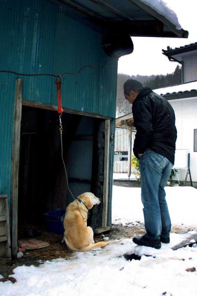 ひとりで家に残るメス犬に話しかける平山さん。「寒いだろう」と、自分の車の助手席に乗せていた座布団を、布団変わりのわらの上に敷いてやった。ここからは、灯りのつかない家と、雪が積もった山しか見えない。車もめったにそばを通らないという。